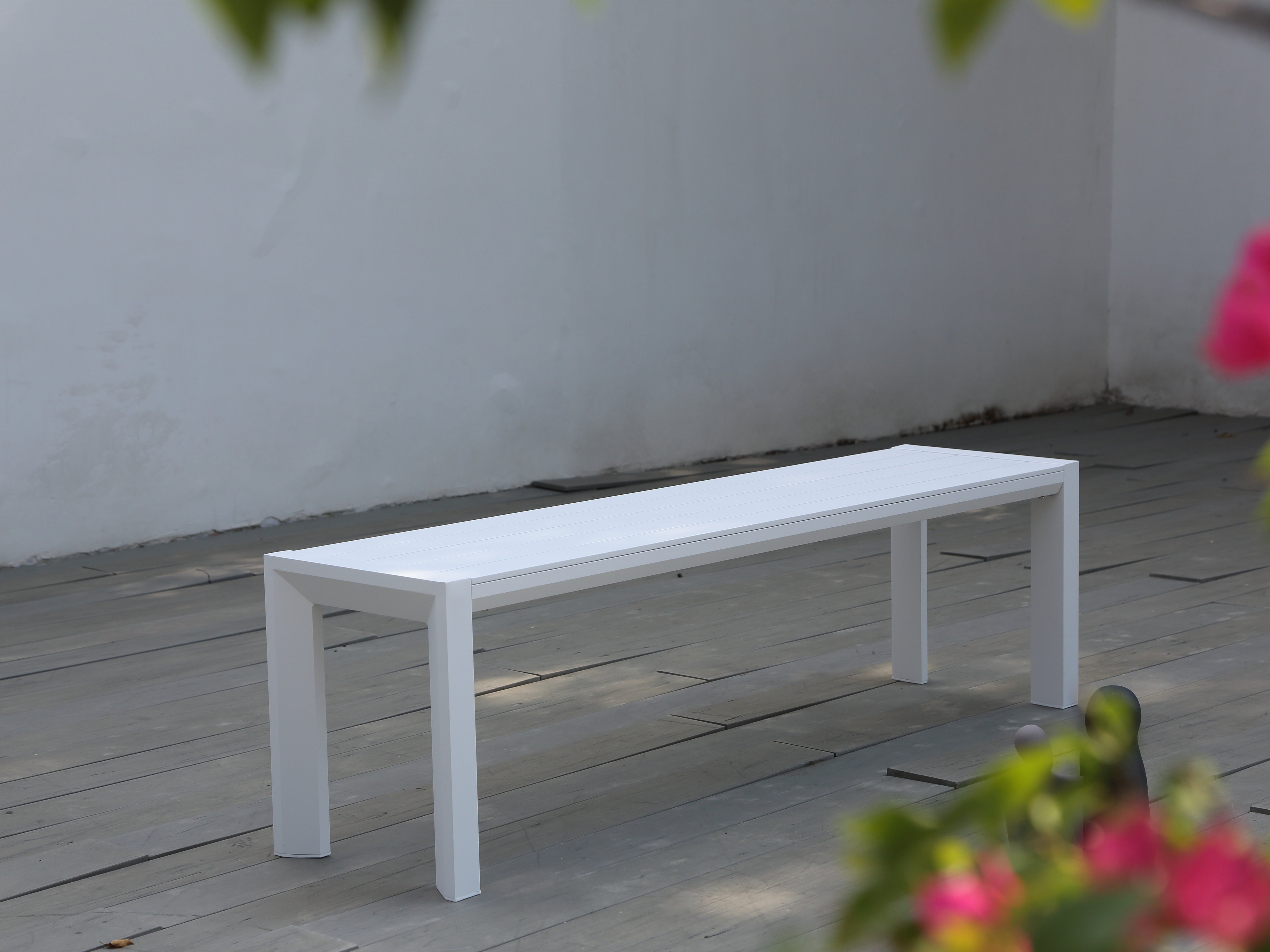 A white aluminium outdoor bench placed on a wooden deck with a wall and greenery in the background.
