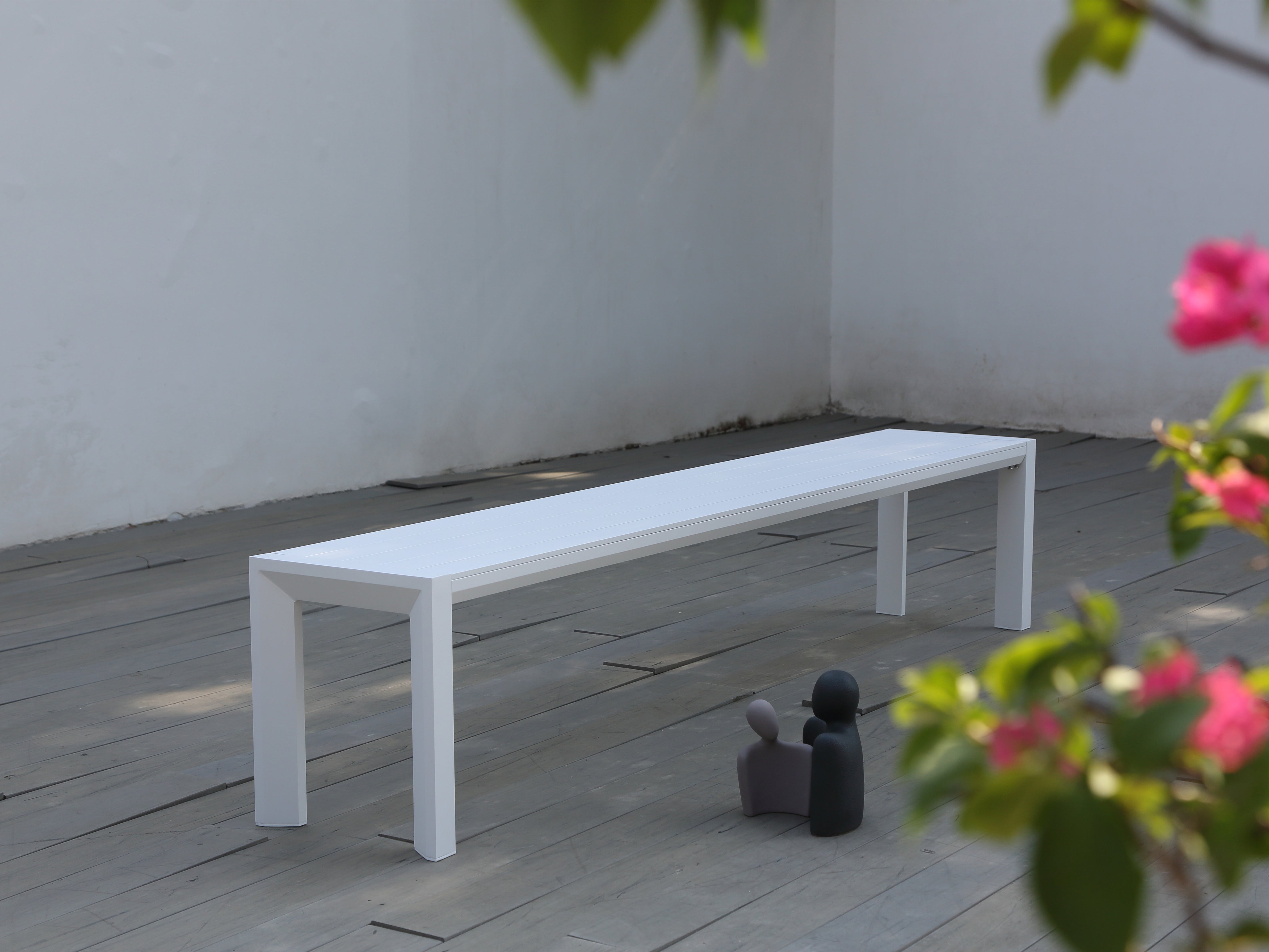 A white aluminium outdoor bench placed on a paved area with a wall in the background and flowers in the foreground.