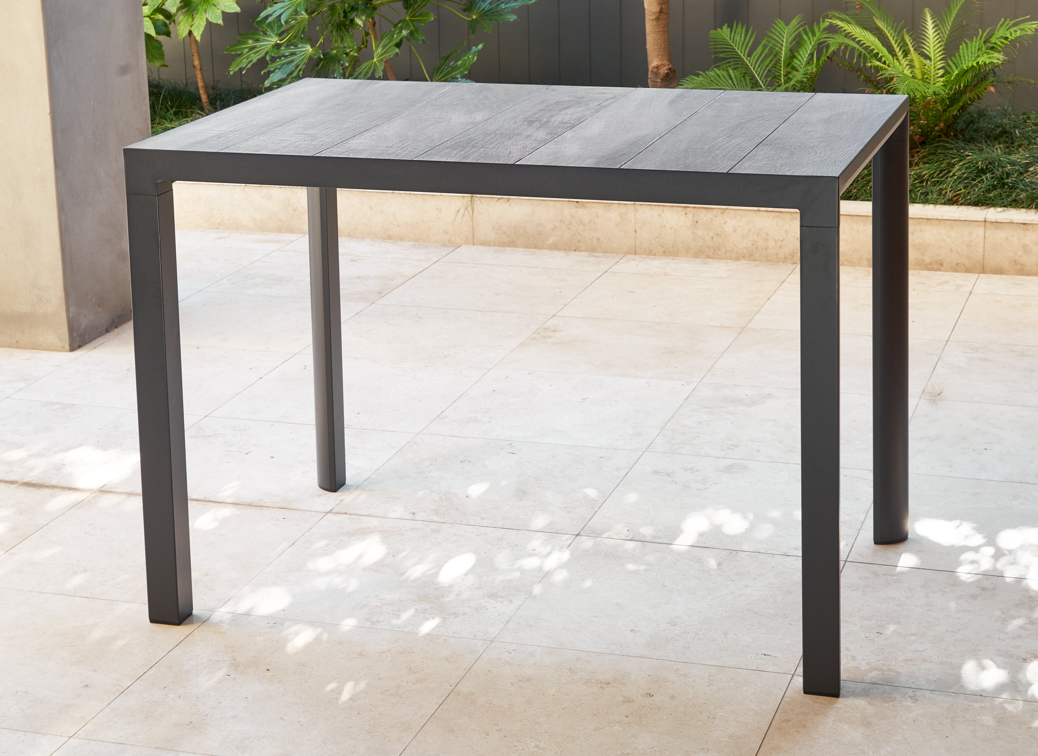 Gray Mayfair Bar Table on a tiled patio with plants in the background